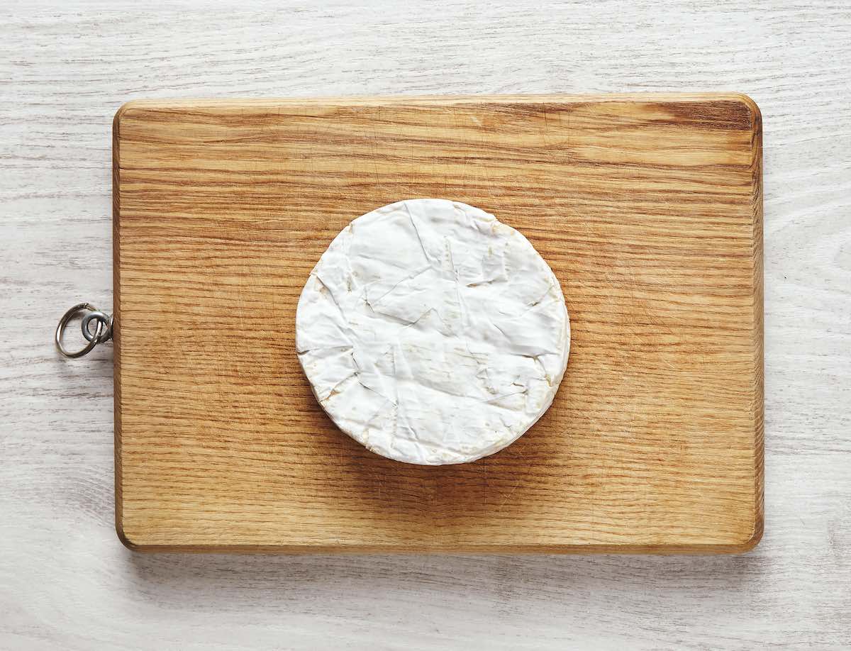 Top View Of Perfect Circle Of Camembert Cheese On Rustic Wooden Board Isolated On Aged White Wooden Table In Center